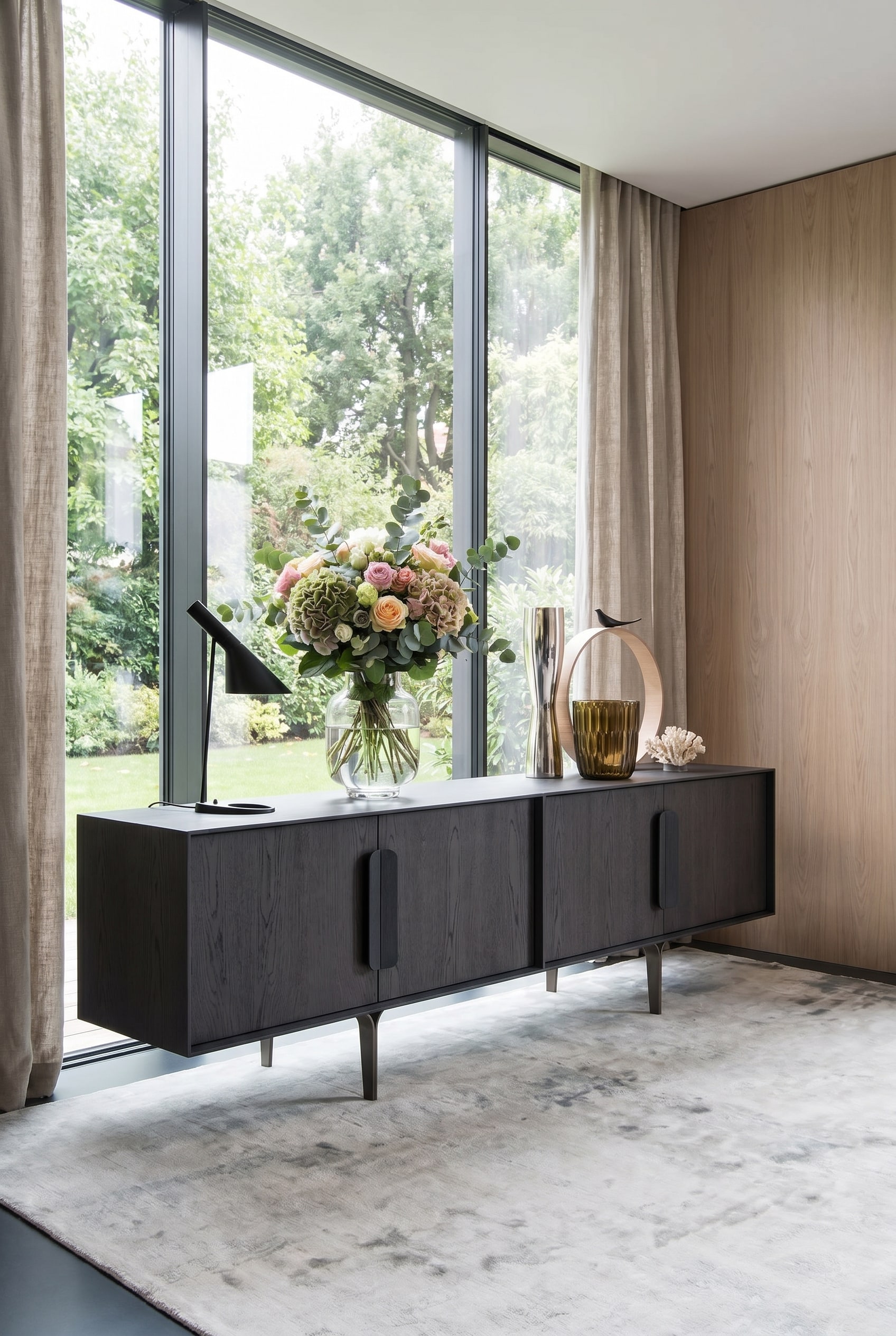 Buffet design foncé devant de grandes fenêtres, sur lequel repose un vase statement lumineux et clair avec des fleurs printanières Buffet design foncé devant de grandes fenêtres, sur lequel repose un vase statement lumineux et clair avec des fleurs printanières