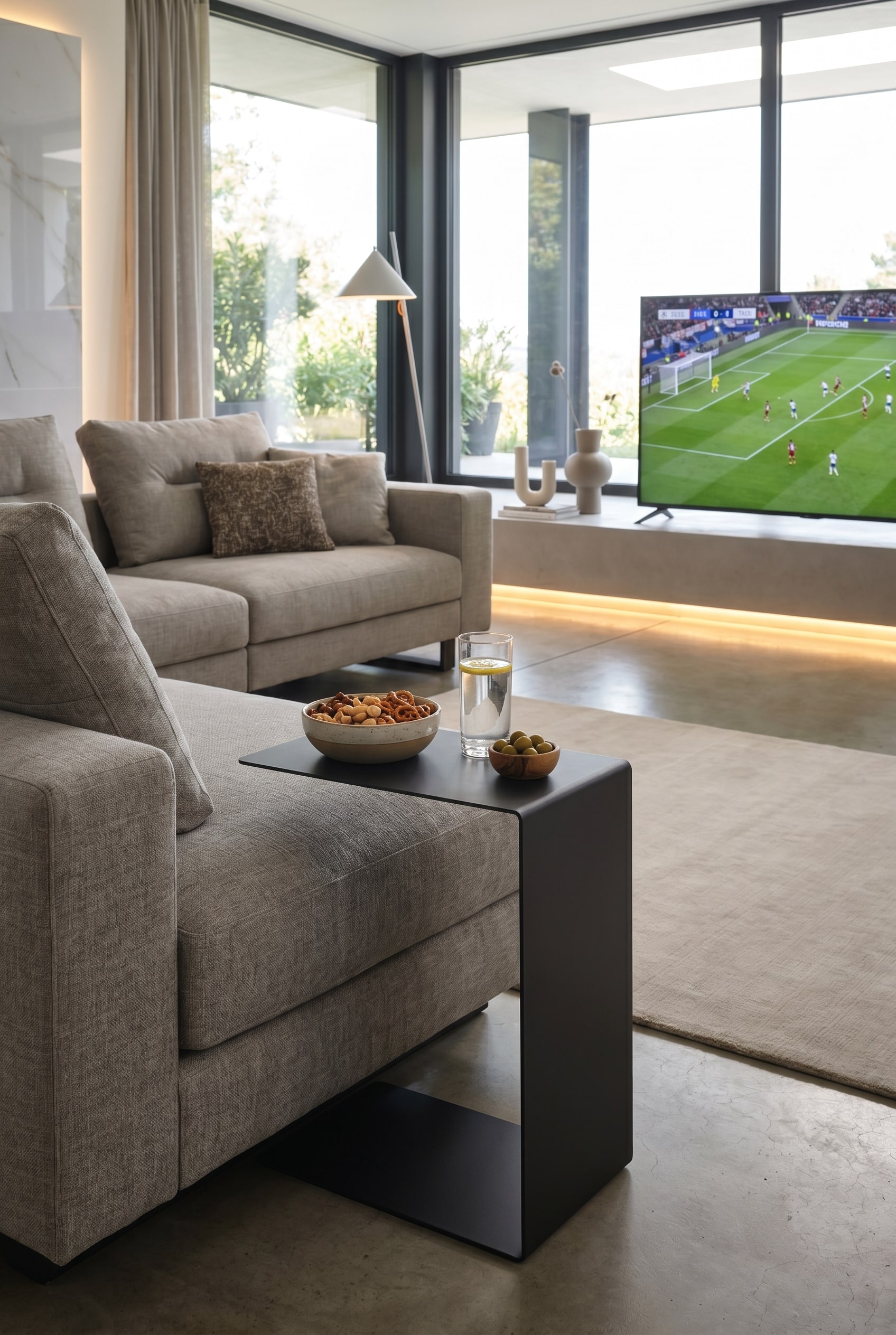 Une table d'appoint moderne Marelli Mini Sofa en forme de C, dont la surface de rangement avec une boisson et des snacks s'&eacute;tend sur l'accoudoir d'un canap&eacute; confortable.