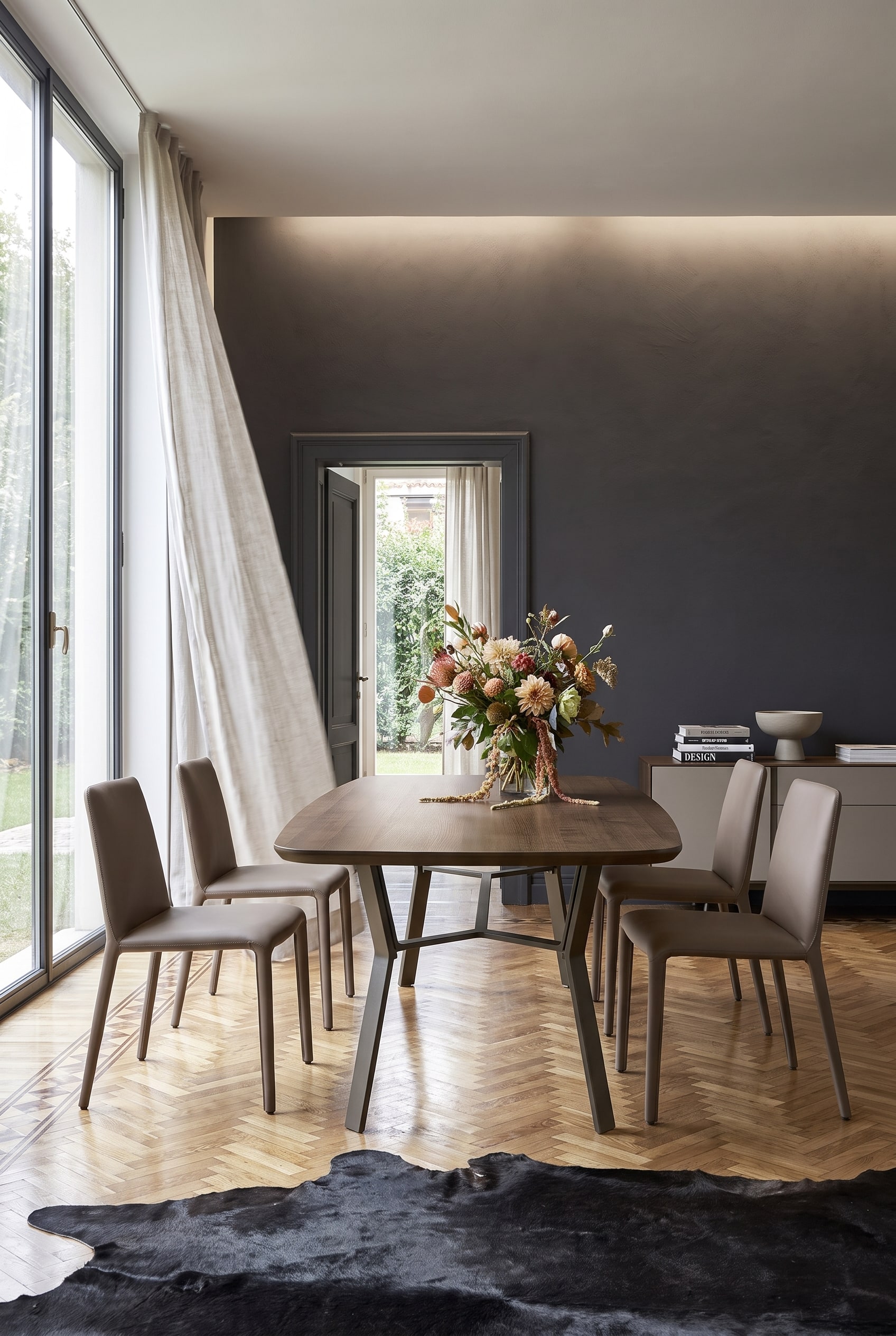 Salle à manger minimaliste avec mur d'accent sombre, table en bois sur piètement en fer et quatre chaises en cuir couleur taupe