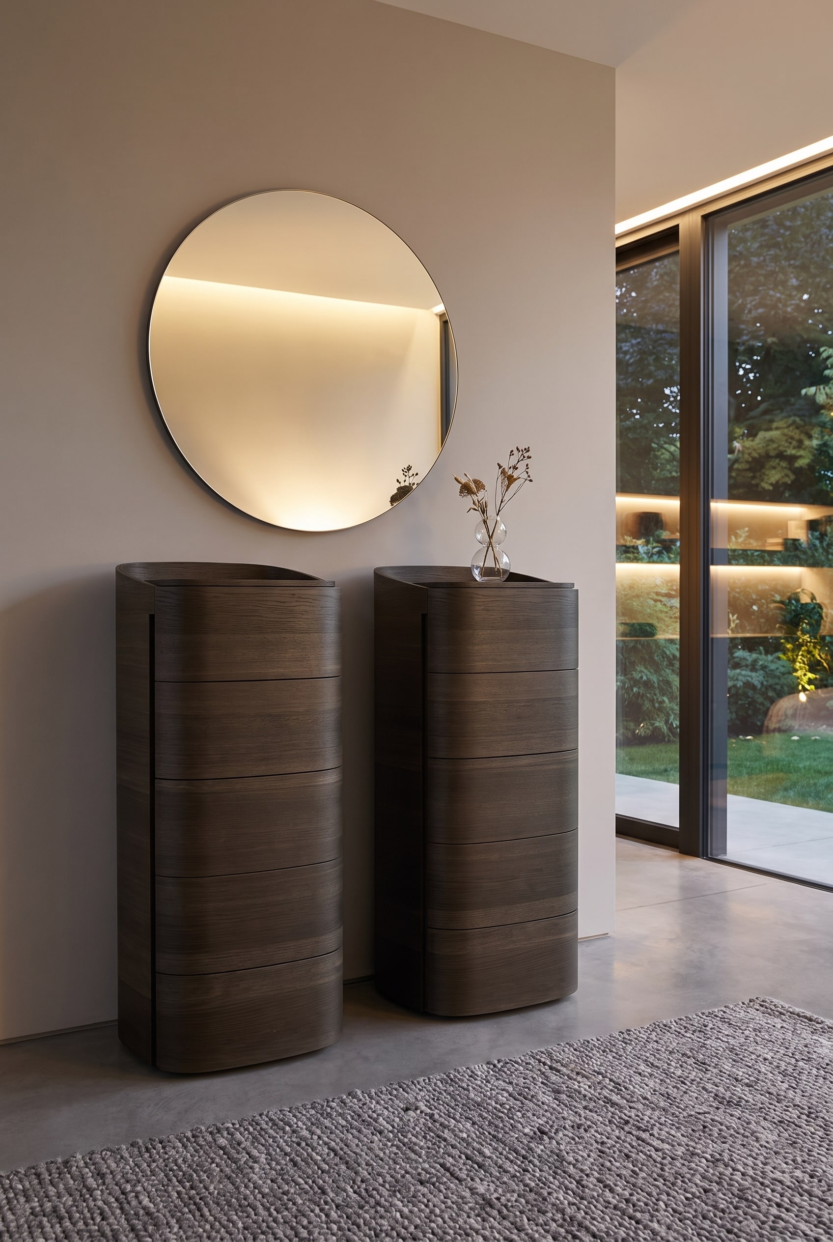 Commode haute et arrondie en bois foncé, surmontée d'un grand miroir rond minimaliste au mur, avec un petit vase devant