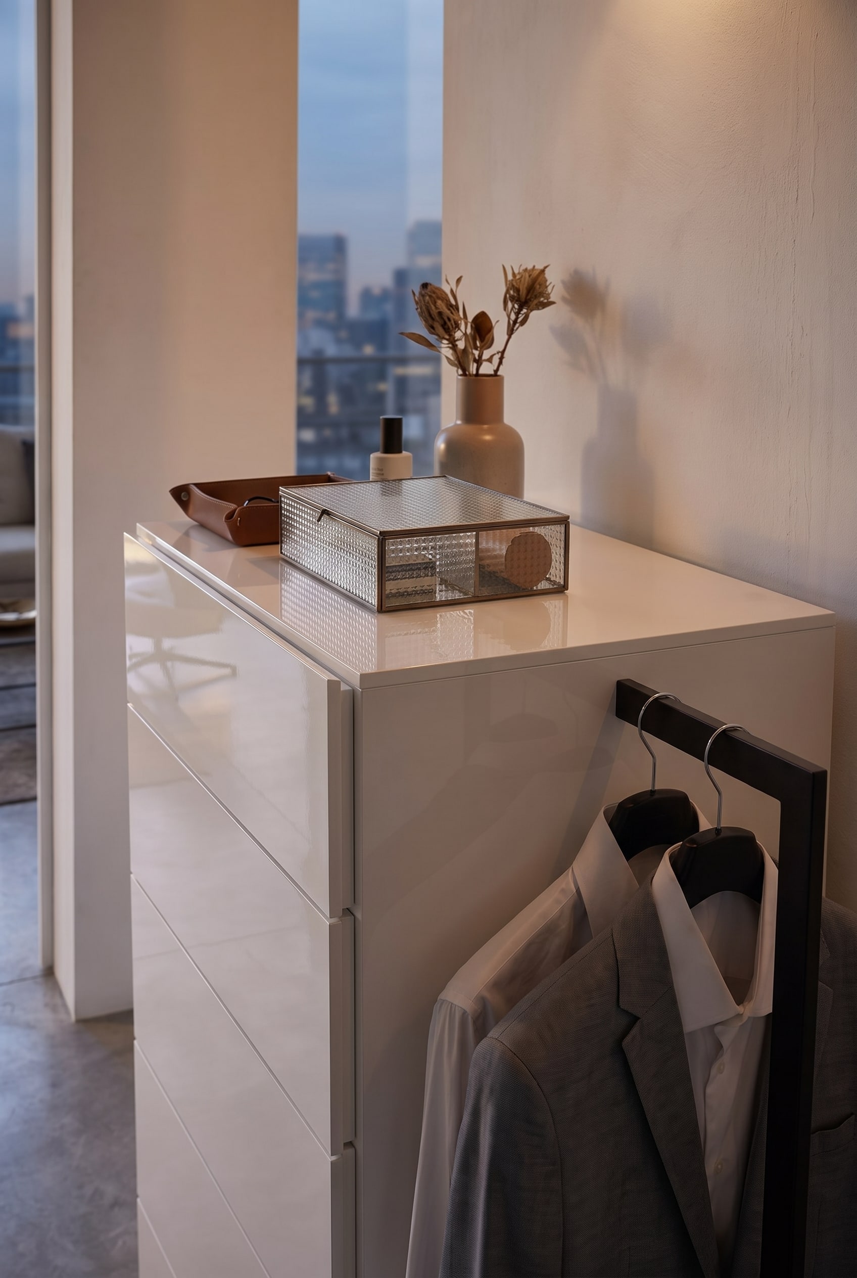 Vue détaillée d'une commode blanche avec une boîte en verre cannelé, un plateau en cuir et une tringle à vêtements latérale avec une veste grise.