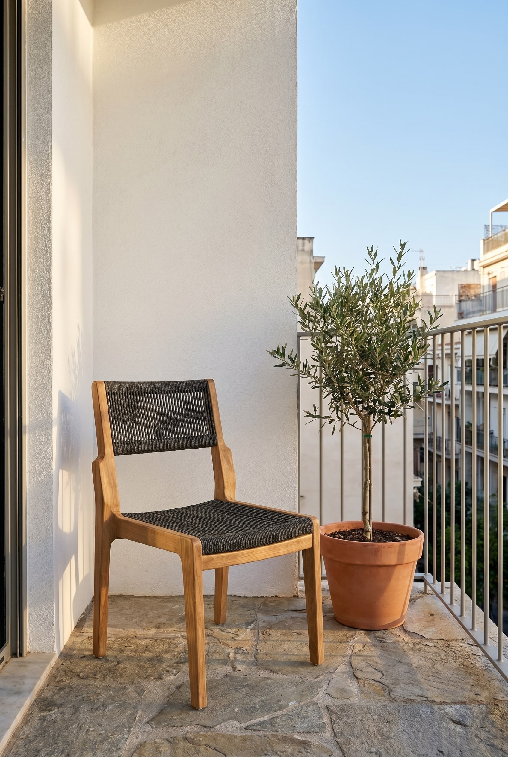 Balcon compact avec sol en pierre naturelle et une chaise en teck avec tressage en corde anthracite sous la douce lumi&egrave;re du matin