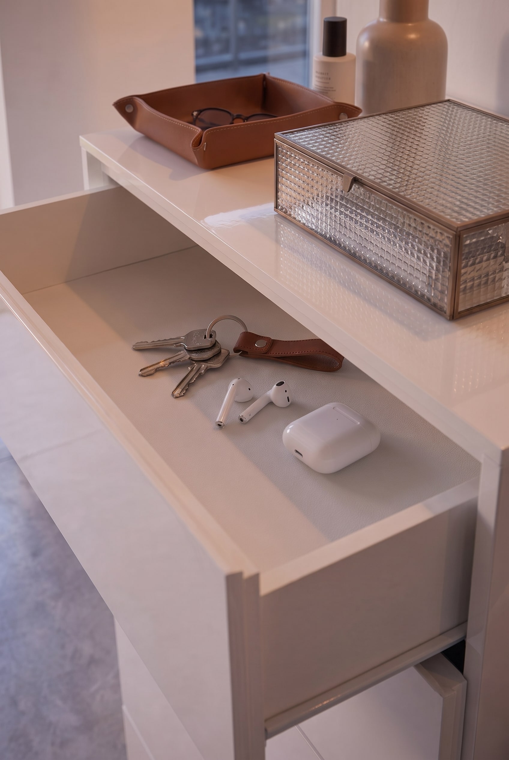 Tiroir plat ouvert d'une commode blanche, contenant des clés, des AirPods et un porte-clés en cuir rangés.