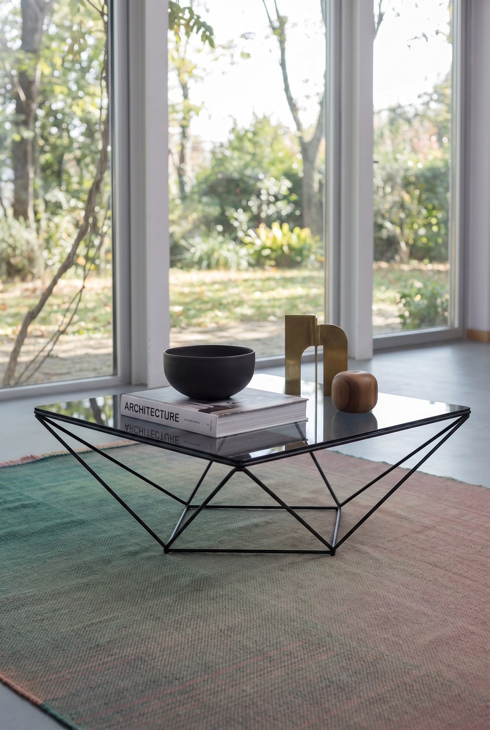 Salon aéré avec table basse carrée en verre et cadre métallique fin, sur laquelle se trouvent un livre d'art architectural et une coupelle en céramique foncée