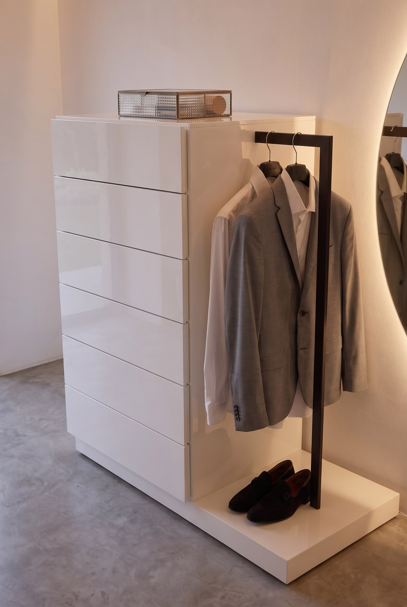 Commode blanche avec tringle de penderie noire lat&eacute;rale. Une veste grise est suspendue au-dessus d'une paire de chaussures fonc&eacute;es sur la plinthe.