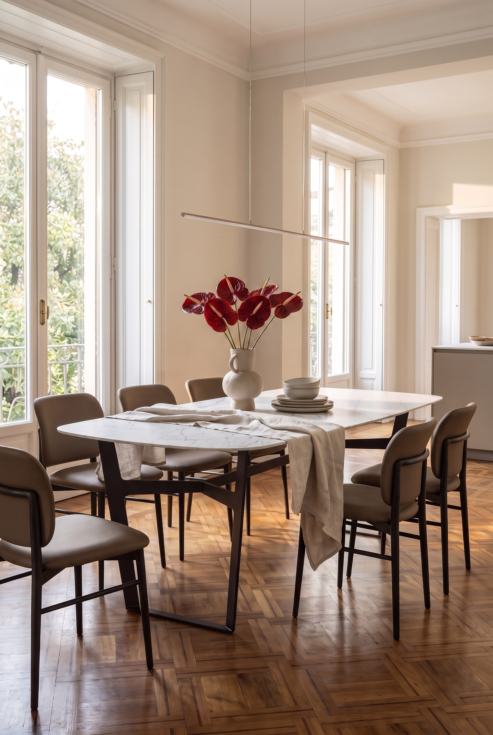 Coin repas lumineux avec chaises en bois fonc&eacute;. Sur la table &agrave; manger en marbre, un vase avec des anthuriums rouge fonc&eacute; est pos&eacute; &agrave; c&ocirc;t&eacute; de vaisselle empil&eacute;e.