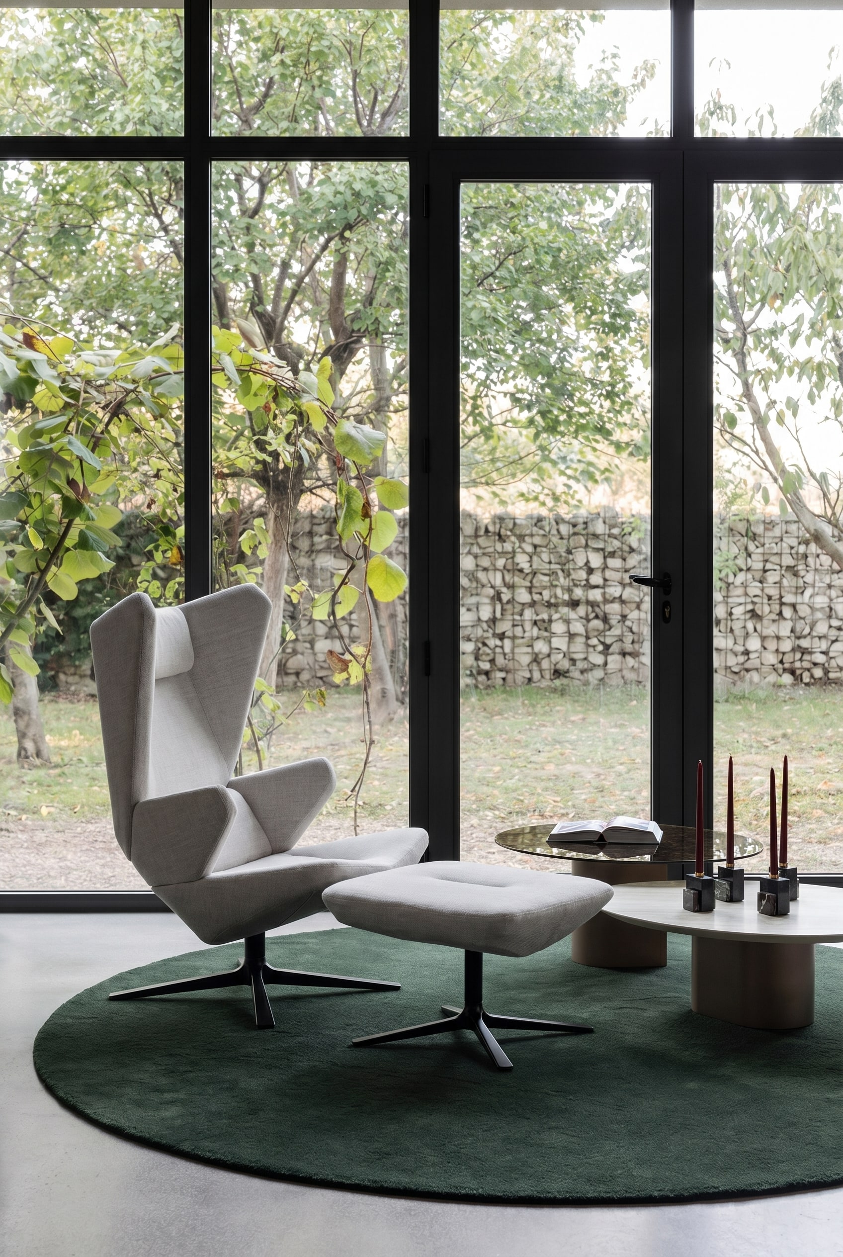 Fauteuil sculptural gris à dossier haut sur un tapis rond vert foncé dans un salon minimaliste avec de grandes fenêtres