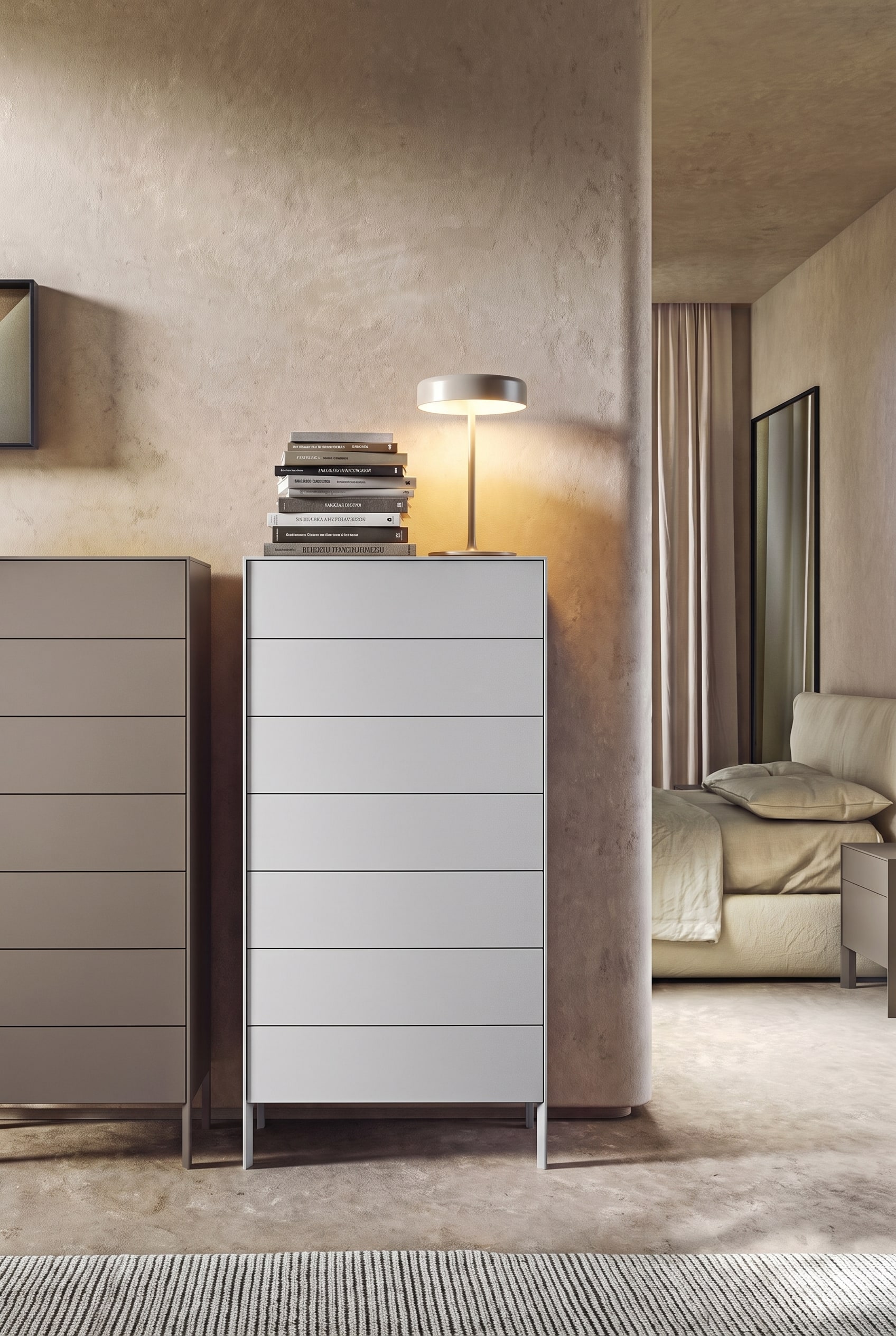 Chambre minimaliste avec une commode grise haute, sur le côté gauche une pile massive de livres et une lampe de table, le côté droit vide